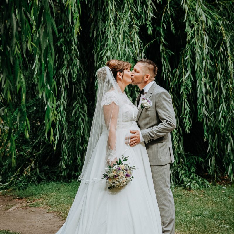Hochzeitsbild von schwangerer Braut küsst ihren Ehemann unter Weidenbäumen im Stadtpark in Bernau